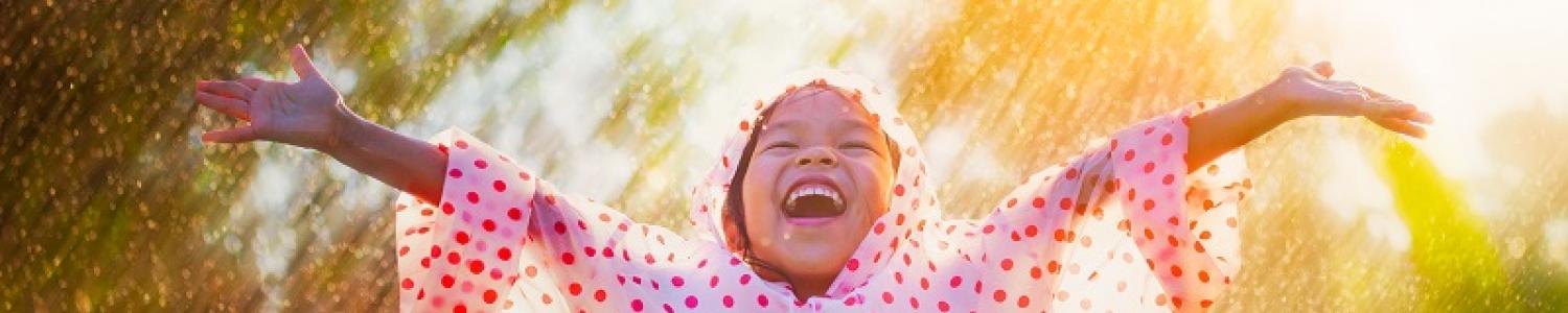 Child in a spotted raincoat with arms joyously outstretched up to the sky