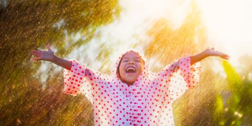 Child in a spotted raincoat with arms joyously outstretched up to the sky